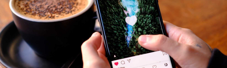 Person holding phone engaging with a picture on Instagram. Coffee in the background.